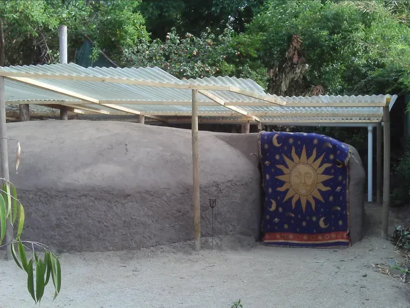 La cúpula de barro sin encalar, de color tierra, bajo un techo de planchas onduladas sostenido por postes de madera. Un textil azul con un sol dorado bordado cubre la entrada.