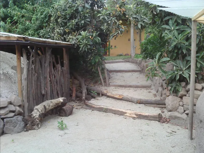 Una escalera de tierra con peldaños contenidos por troncos, entre un cobertizo de postes de madera y un muro de piedras sueltas, bajo árboles frutales; al fondo, una casa de muros amarillos.