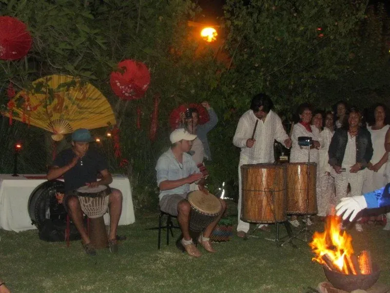 De noche, en el jardín, dos músicos sentados tocan tambores de mano y otro golpea de pie un tambor grande; un grupo de asistentes vestidos de blanco escucha alrededor y en primer plano arde una fogata.